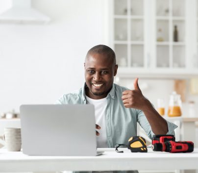 Review of construction instruments. Happy black handyman looking at laptop webcam and showing thumb
